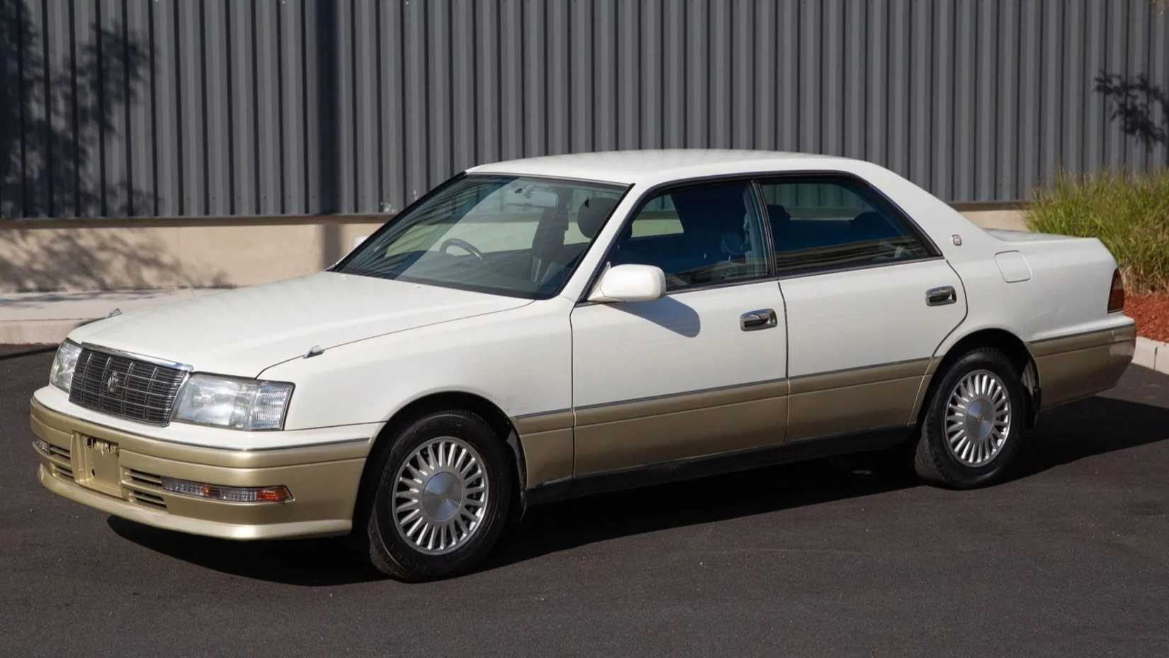 Um Toyota Crown S170 branco e dourado estacionado em frente a um armazém cinzento.