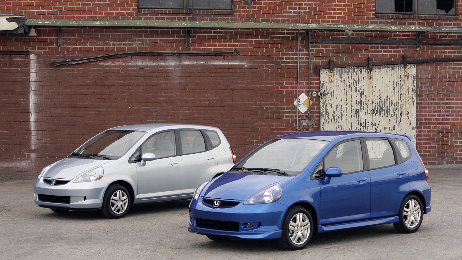 2007 Honda Fit In silver and 2007 Honda Fit Sport in blue Posing in front of wall