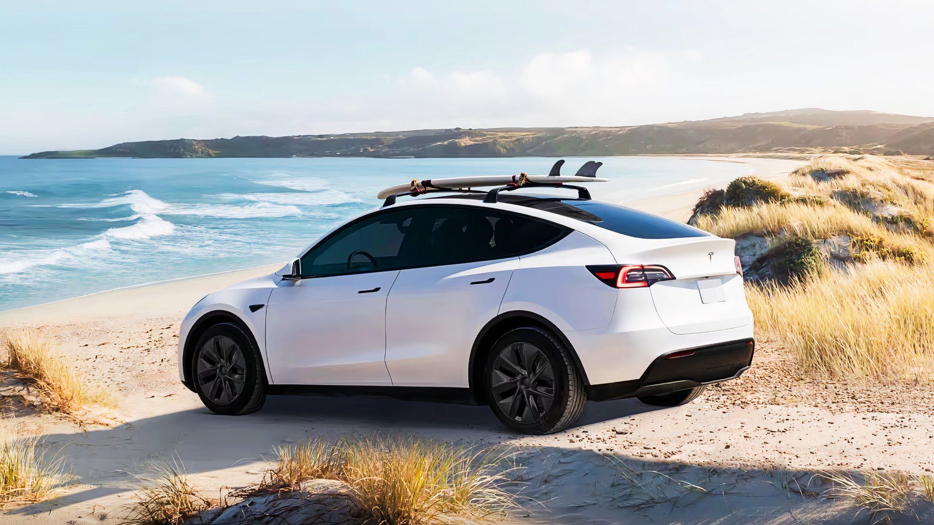 Um pequeno SUV elétrico branco Tesla Model Y 2024 está estacionado em uma praia arenosa.