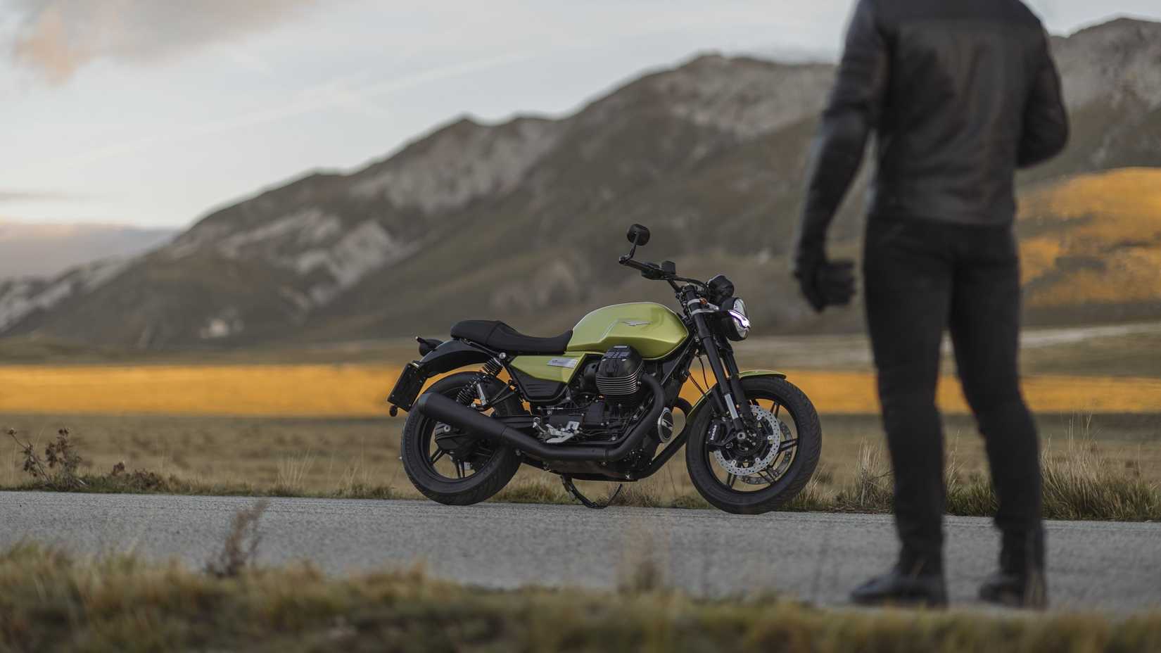 A 2025 Moto Guzzi V7 Sport parked on the side of the road, with the rider in the foreground and fields and mountains in the background.