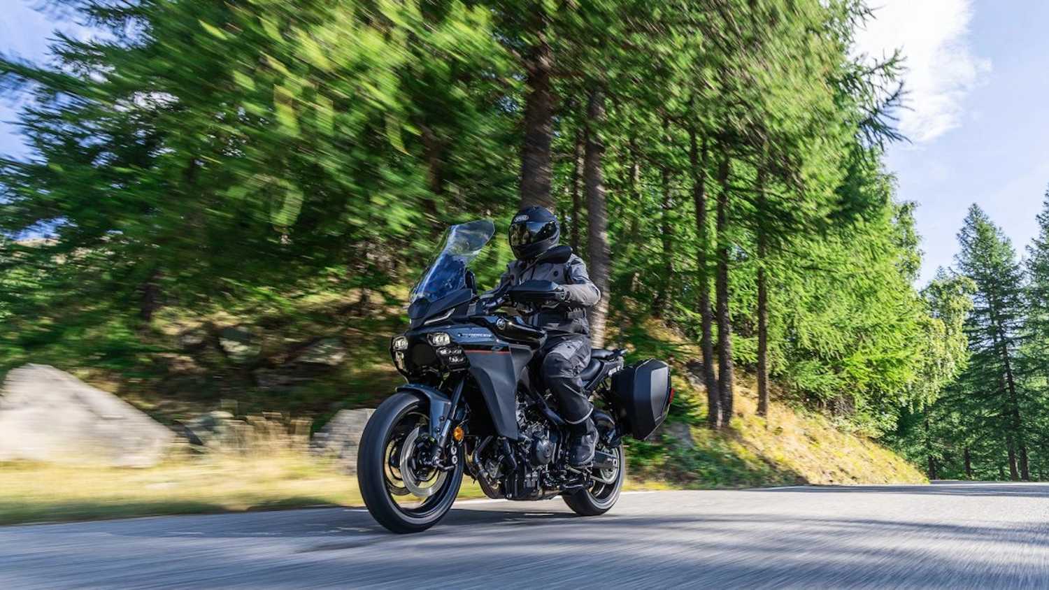 Une Yamaha Tracer 9 équipée de valises latérales, en roulage sur une route de montagne bordée de sapins