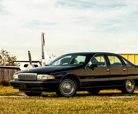 1991 Chevrolet Caprice in black parked on grass in front of airplane