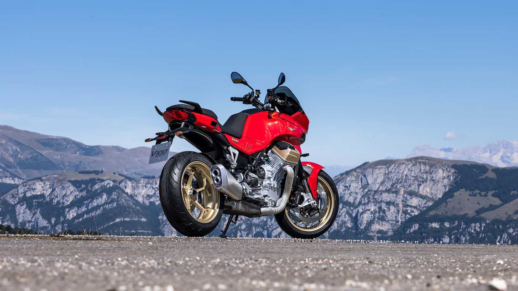 A low rear right three quarter shot of a red Moto Guzzi V100 Mandello parked with mountains in the background