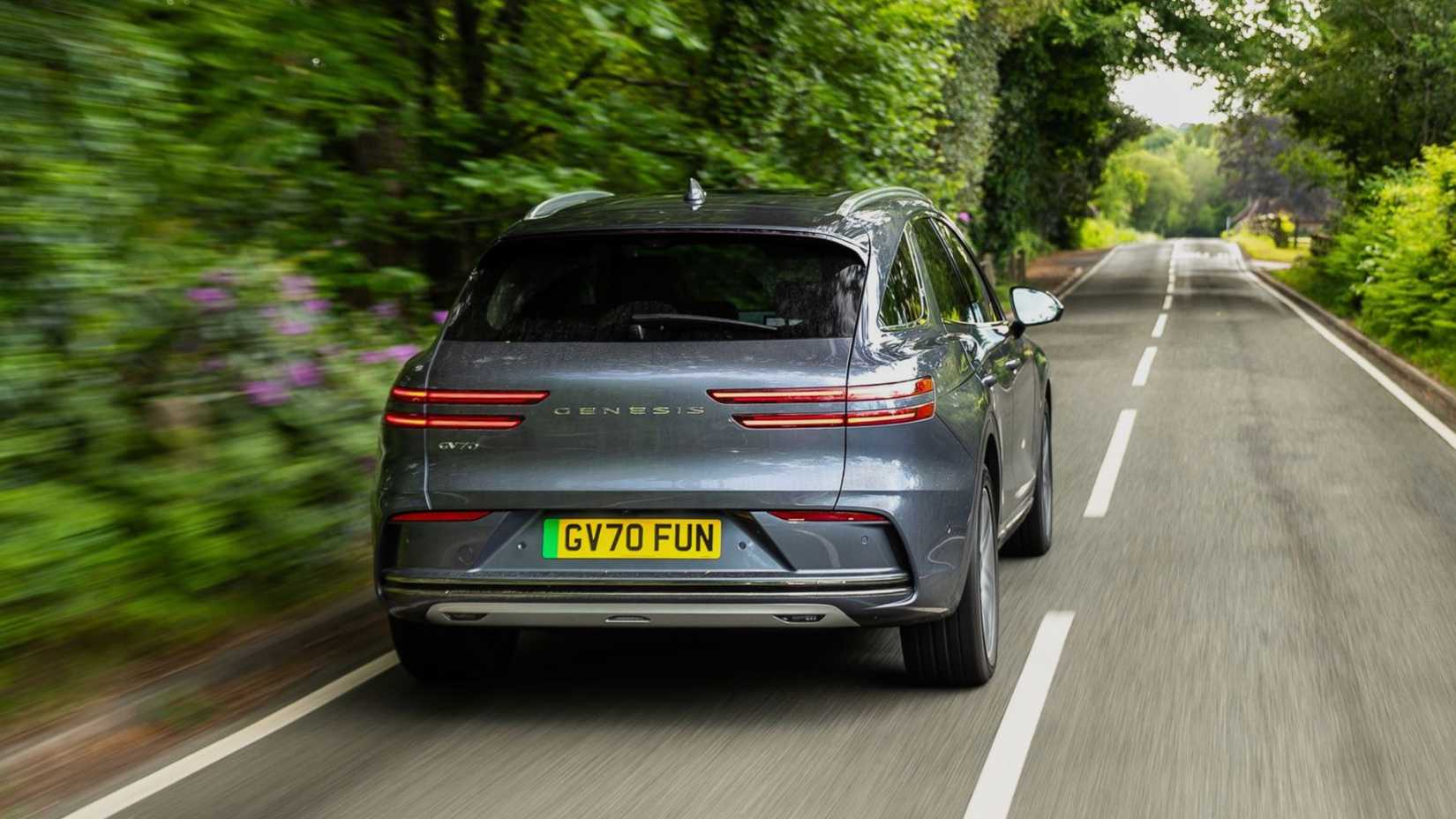 Dynamic rear-end shot of a gray 2026 Genesis Electrified GV70 driving on a country road lined by trees.