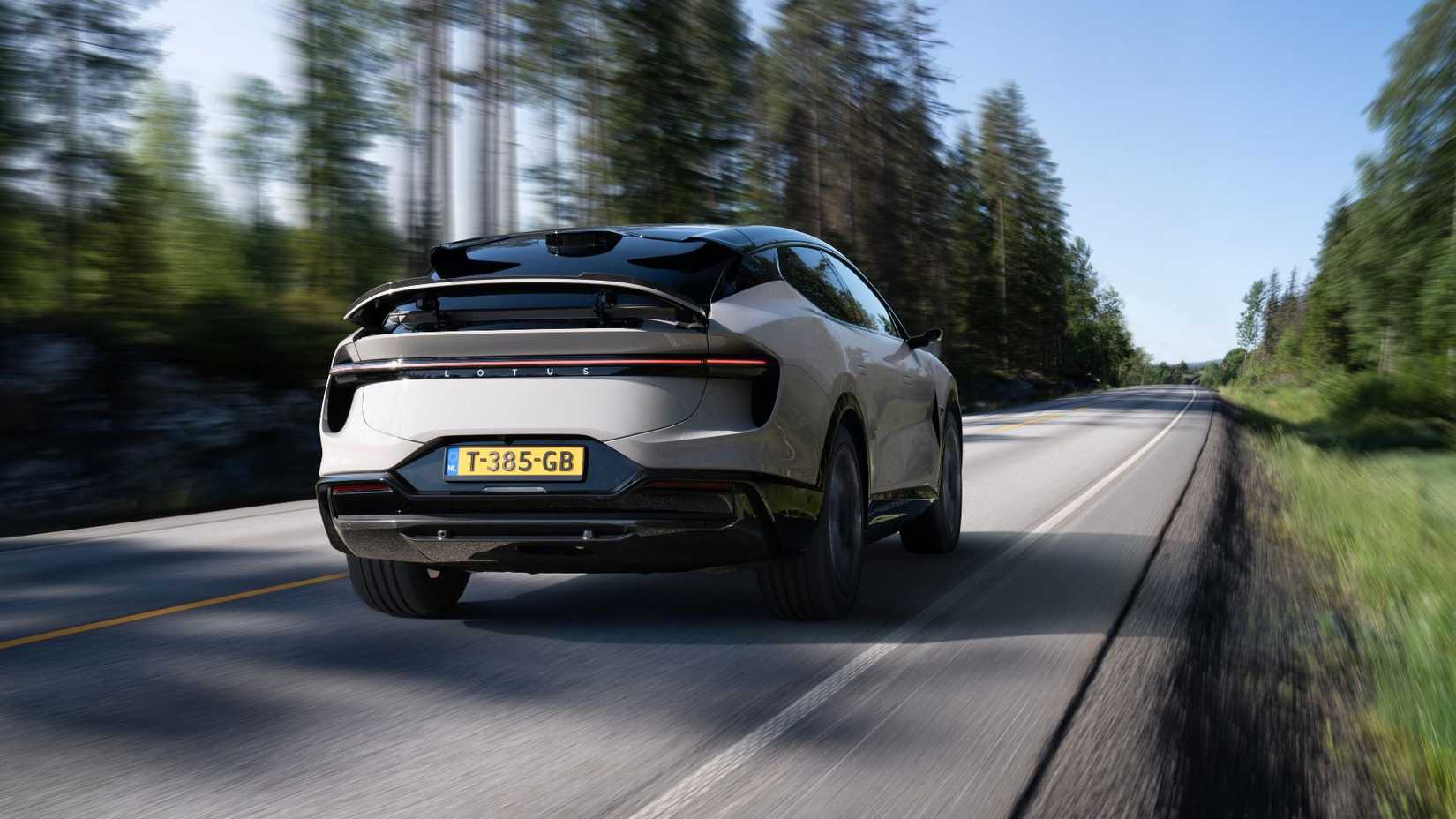 Dynamic rear 3/4 shot of a gray Lotus Eletre S driving on a country road lined by trees.