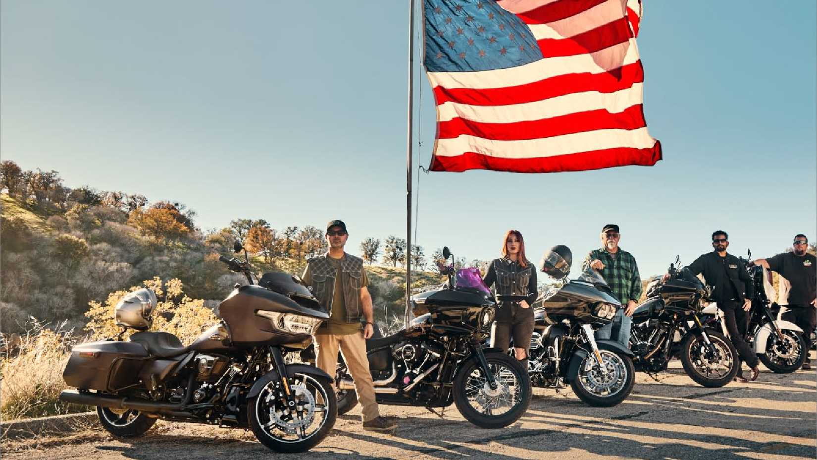 Multiple riders and their Harley motorcycles next to an American flag