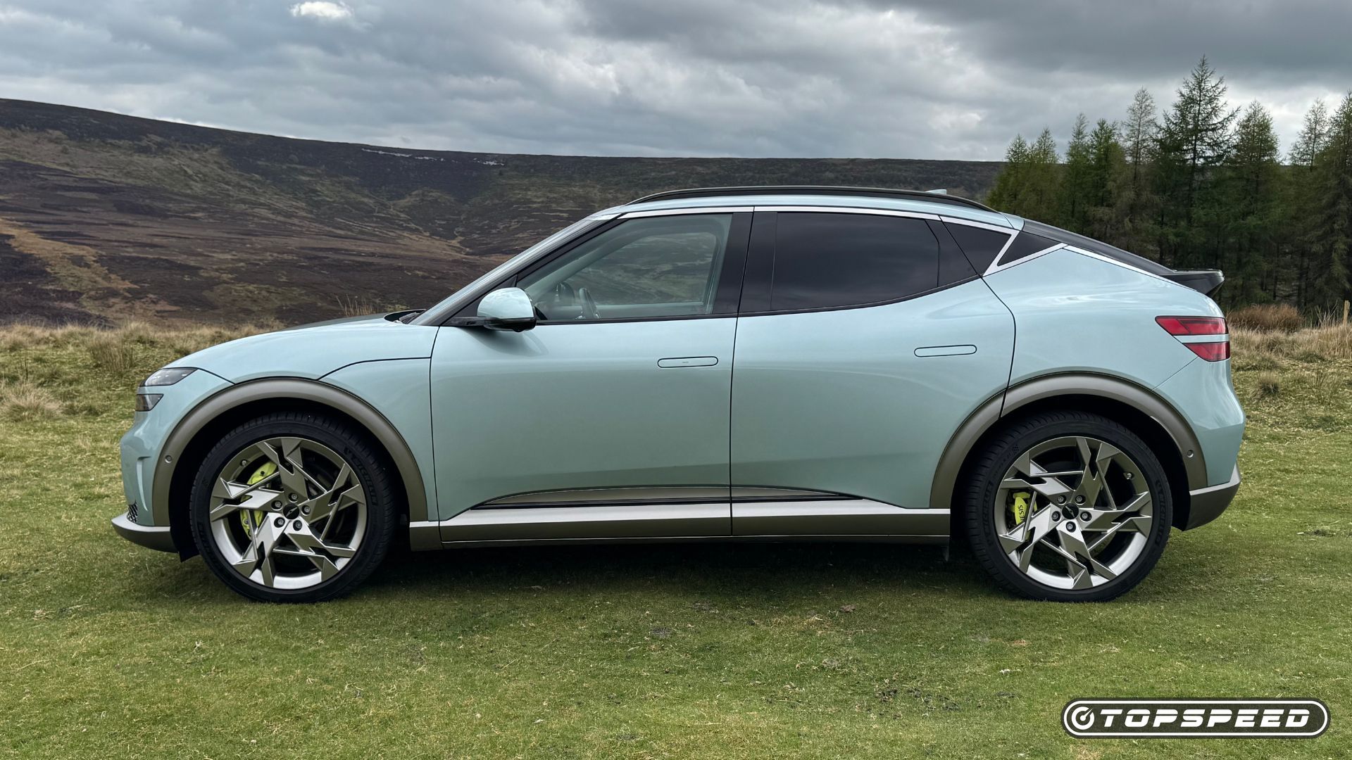 Static side profile shot of a 2024 Genesis GV60 Sport Plus AWD parked on grass with trees in the background.