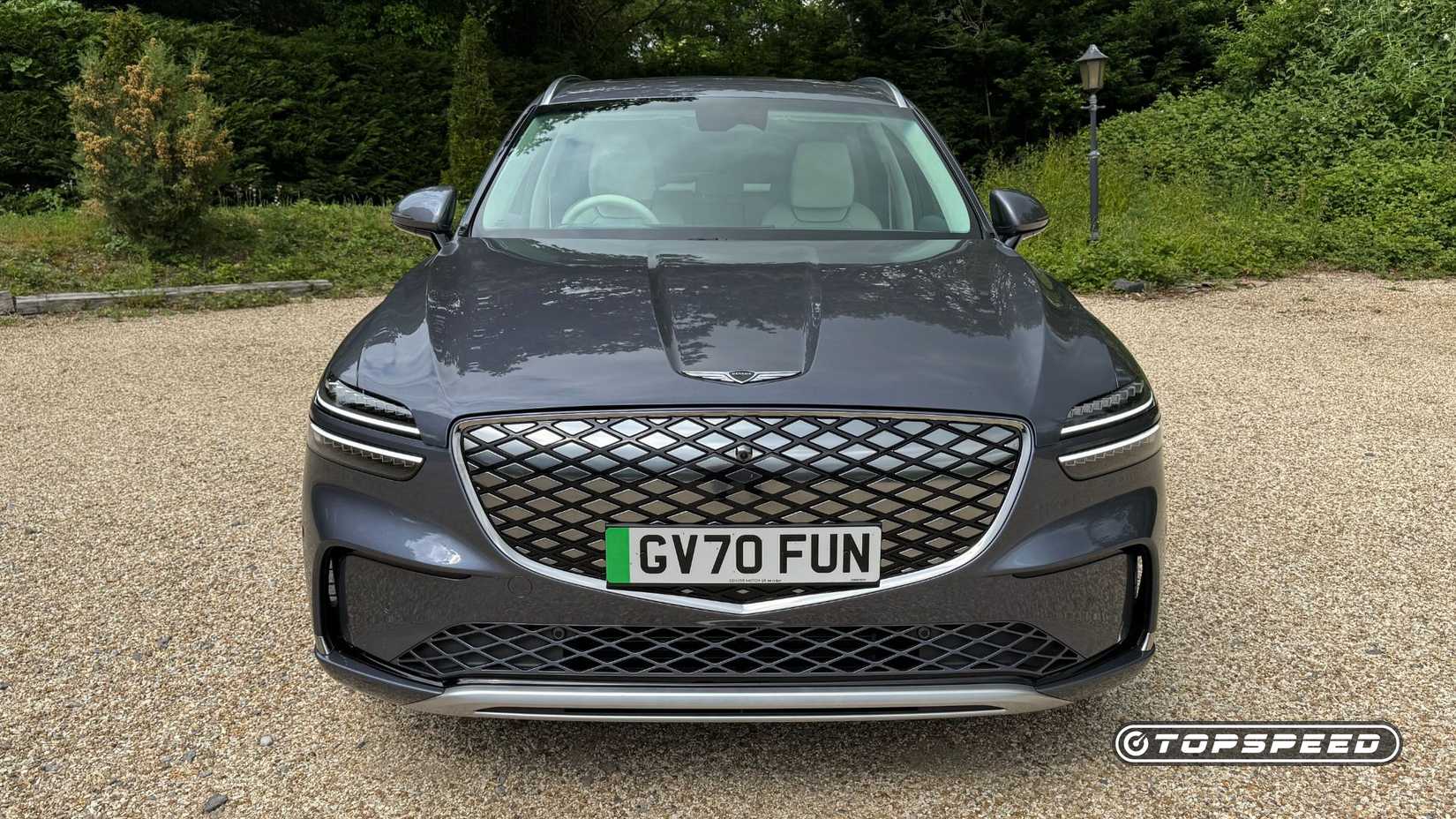 Static front-end shot of a gray 2026 Genesis Electrified GV70 parked on gravel with trees in the background.