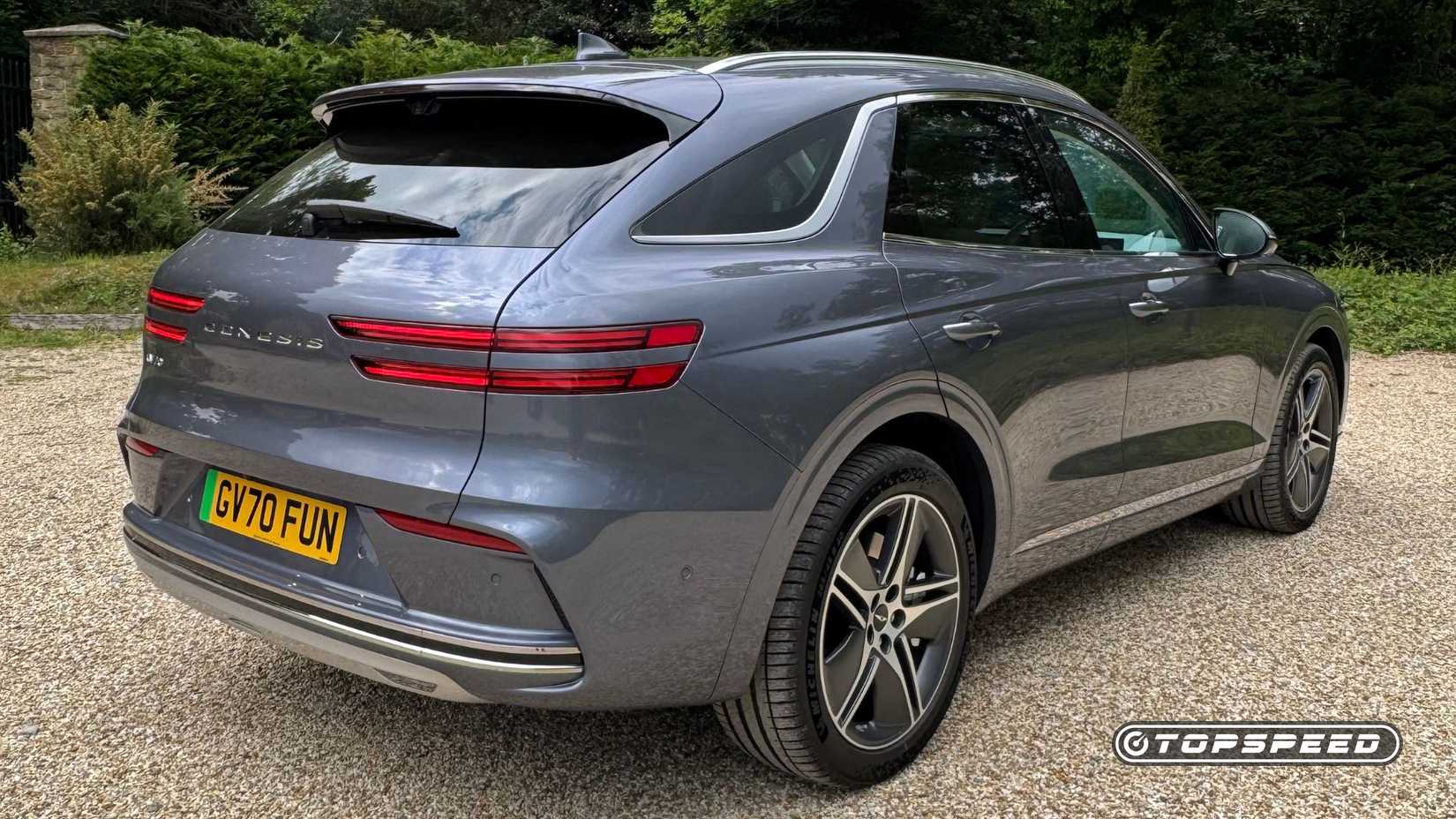 Static rear 3/4 shot of a gray 2026 Genesis Electrified GV70 parked on gravel with trees in the background.
