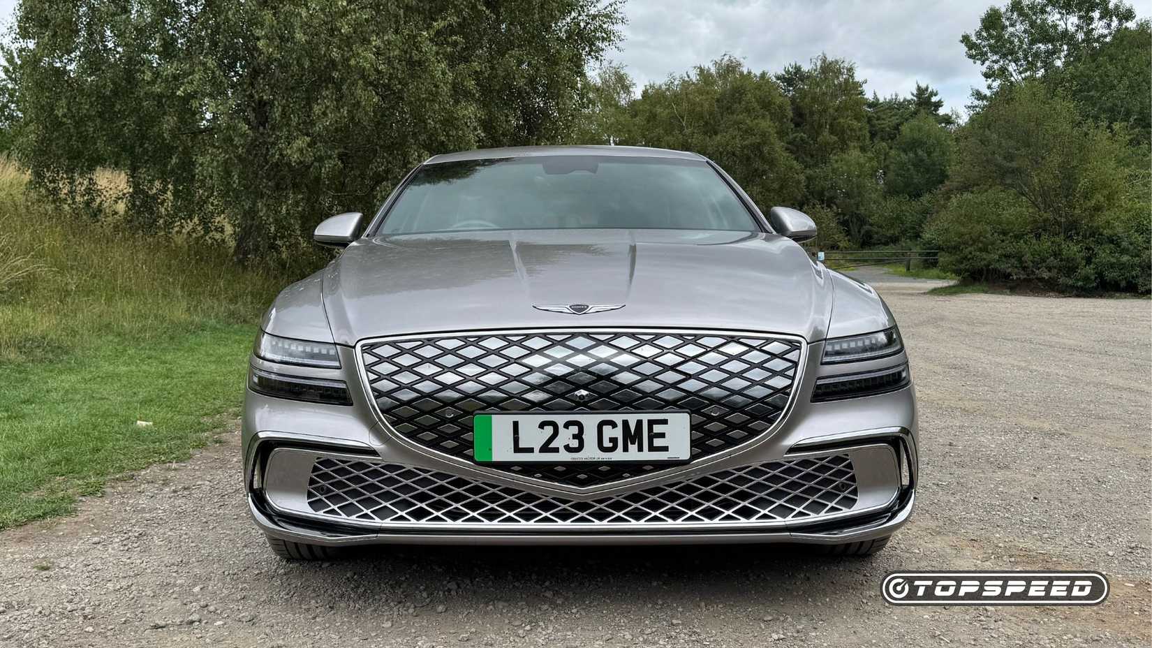 Static front-end shot of a silver 2026 Genesis Electrified G80 parked on gravel with trees in the background.