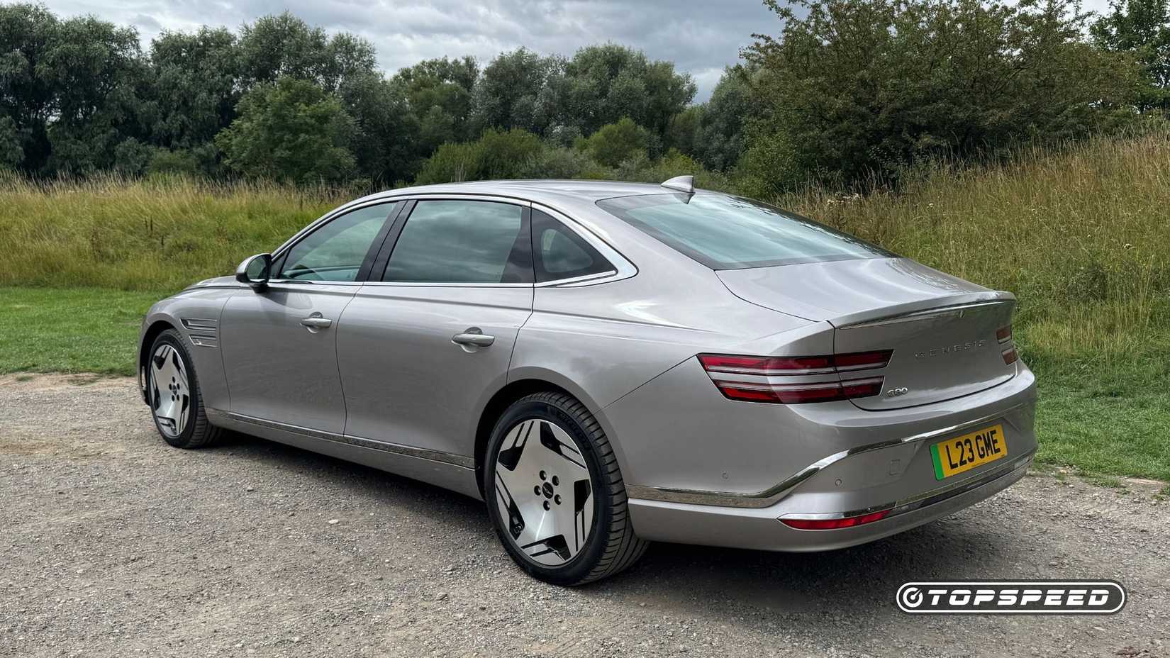 Static rear 3/4 shot of a silver 2026 Genesis Electrified G80 parked on gravel with trees in the background.