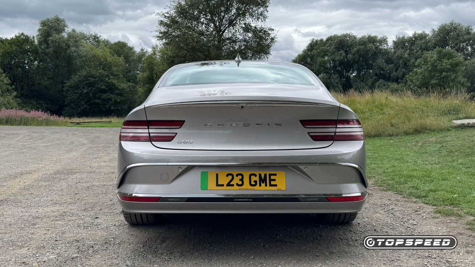 Static rear-end shot of a silver 2026 Genesis Electrified G80 parked on gravel with trees in the background.