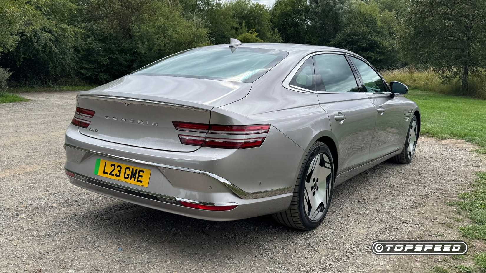 Static rear 3/4 shot of a silver 2026 Genesis Electrified G80 parked on gravel with trees in the background.