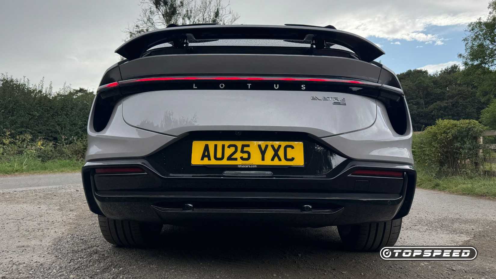 Static rear-end shot of a gray 2025 Lotus Eletre 600 Sport SE with its spoiler deployed wile parked on gravel with trees in the background.