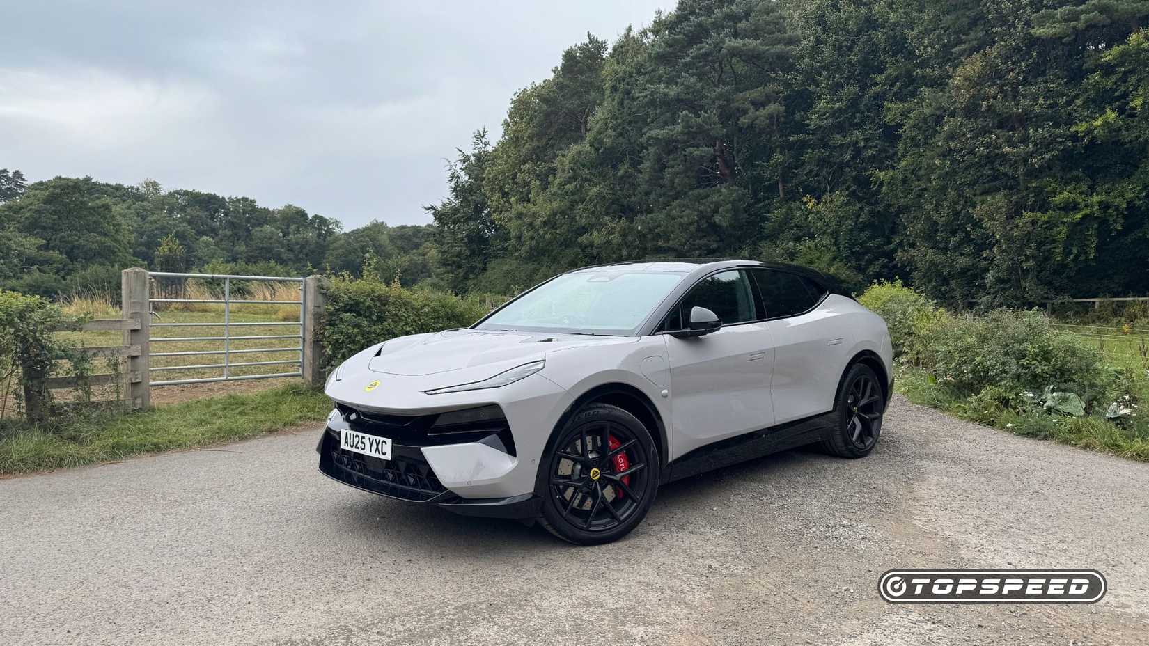 Static front 3/4 shot of a gray 2025 Lotus Eletre 600 Sport SE parked on gravel with trees in the background.